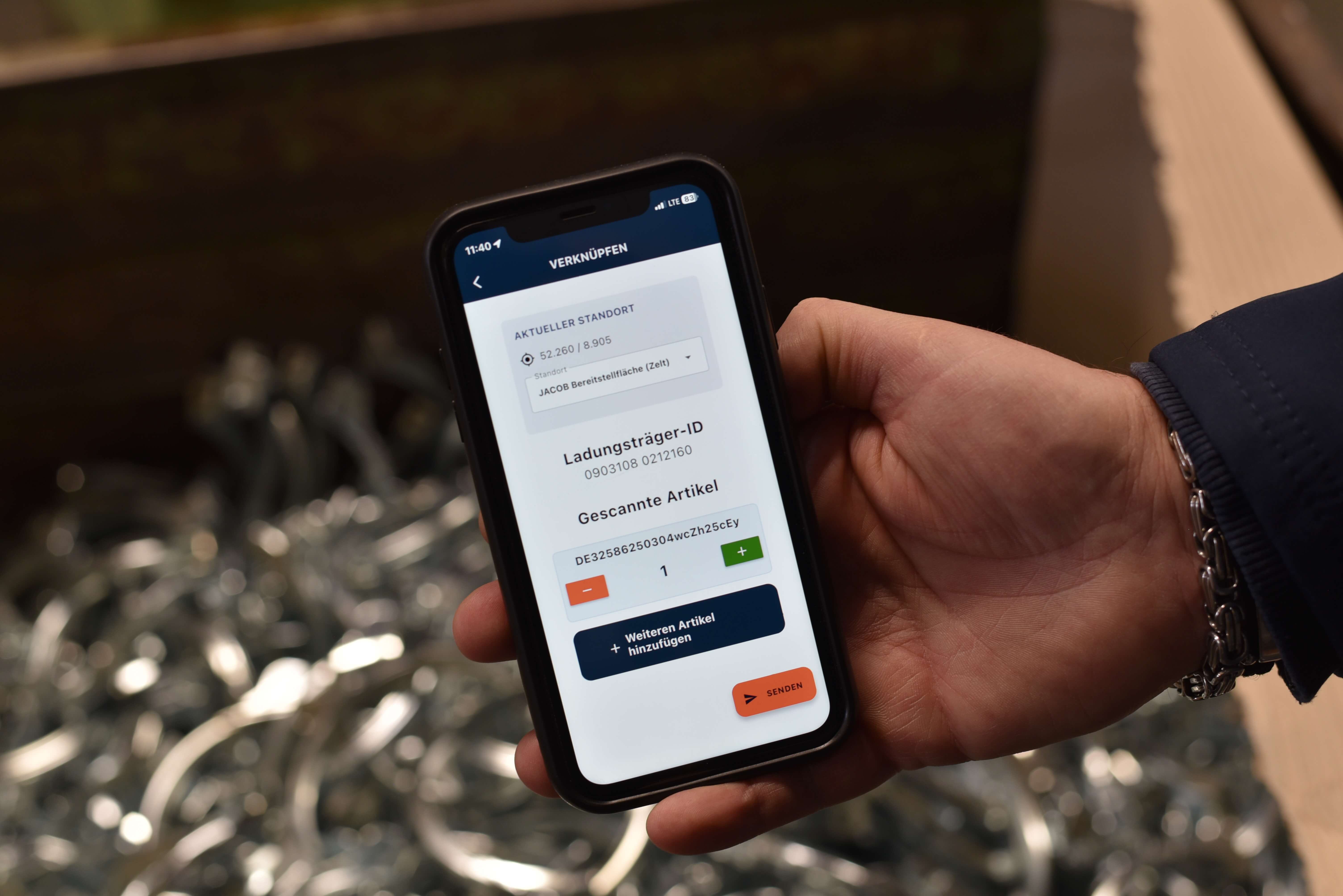 Eine Hand hält ein Smartphone mit der geöffneten Logistikbude-App im „Verknüpfen“-Reiter. Sichtbar sind der aktuelle Standort, der ausgewählte Ladungsträger, der gescannte Artikel samt Menge sowie die Schaltflächen „Weitere Artikel hinzufügen“ und „Senden“.