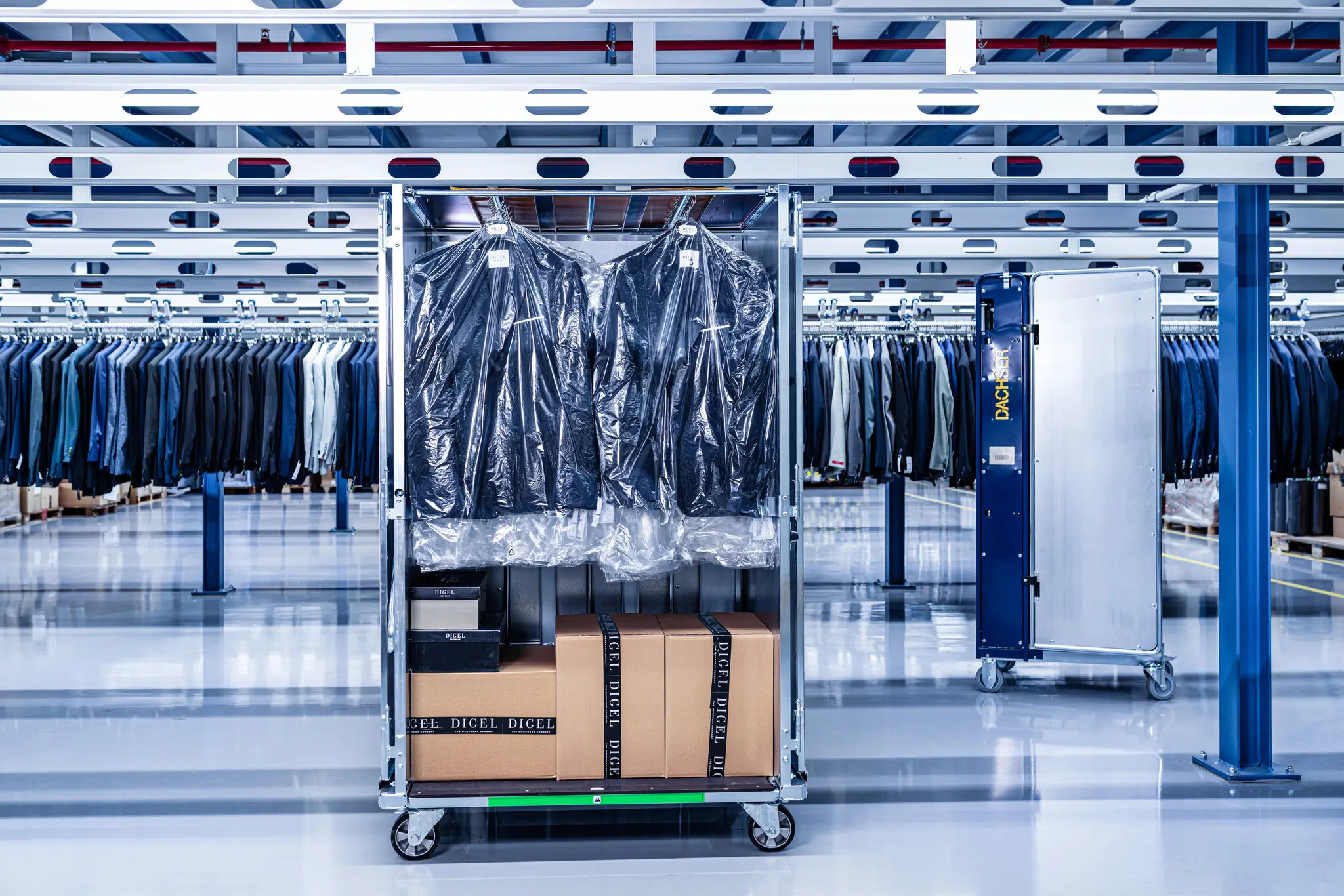 Rollcontainer in einem modernen Logistikzentrum für Bekleidung: Oben hängen Anzüge in transparenten Schutzhüllen, darunter stehen mehrere Kartons mit schwarzem „DIESEL“-Klebeband. Im Hintergrund sind zahlreiche Kleiderstangen mit sortierter Ware sowie eine automatisierte Förderanlage in einer hellen, industriellen Halle zu sehen.