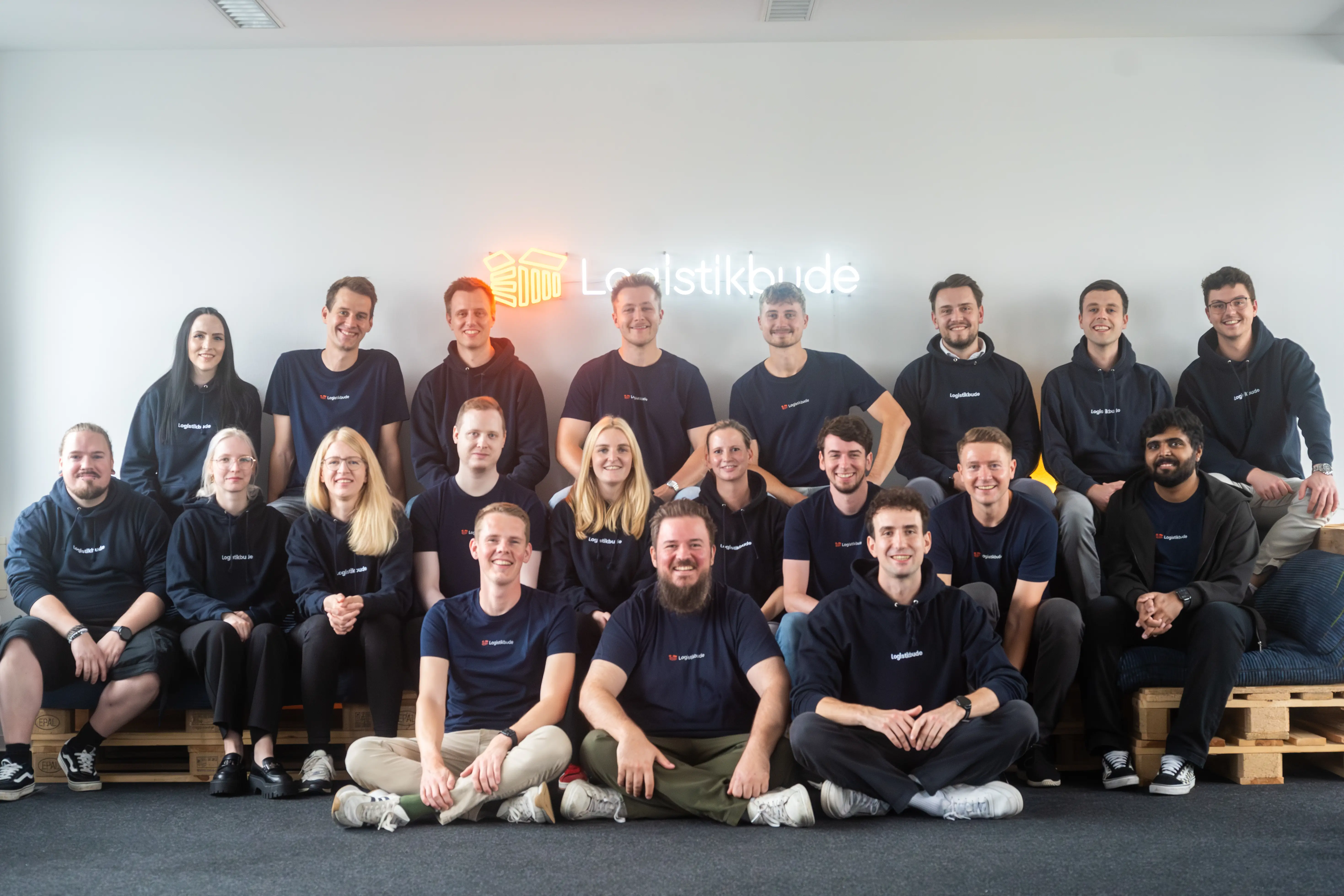 Team photo of Logistikbude: Employees sit and stand together in front of a glowing Logistikbude logo in the office.