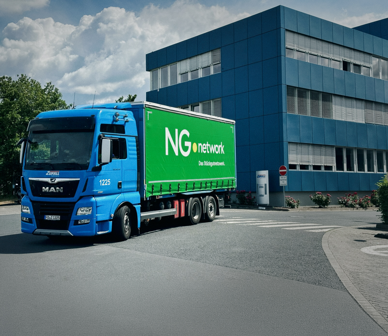 A blue MAN lorry with a green trailer belonging to the haulage firm Zufall is driving around the company premises. The trailer bears the logo and the words “NG network – The General Cargo Network”. In the background, the blue-clad Zufall company building can be seen.