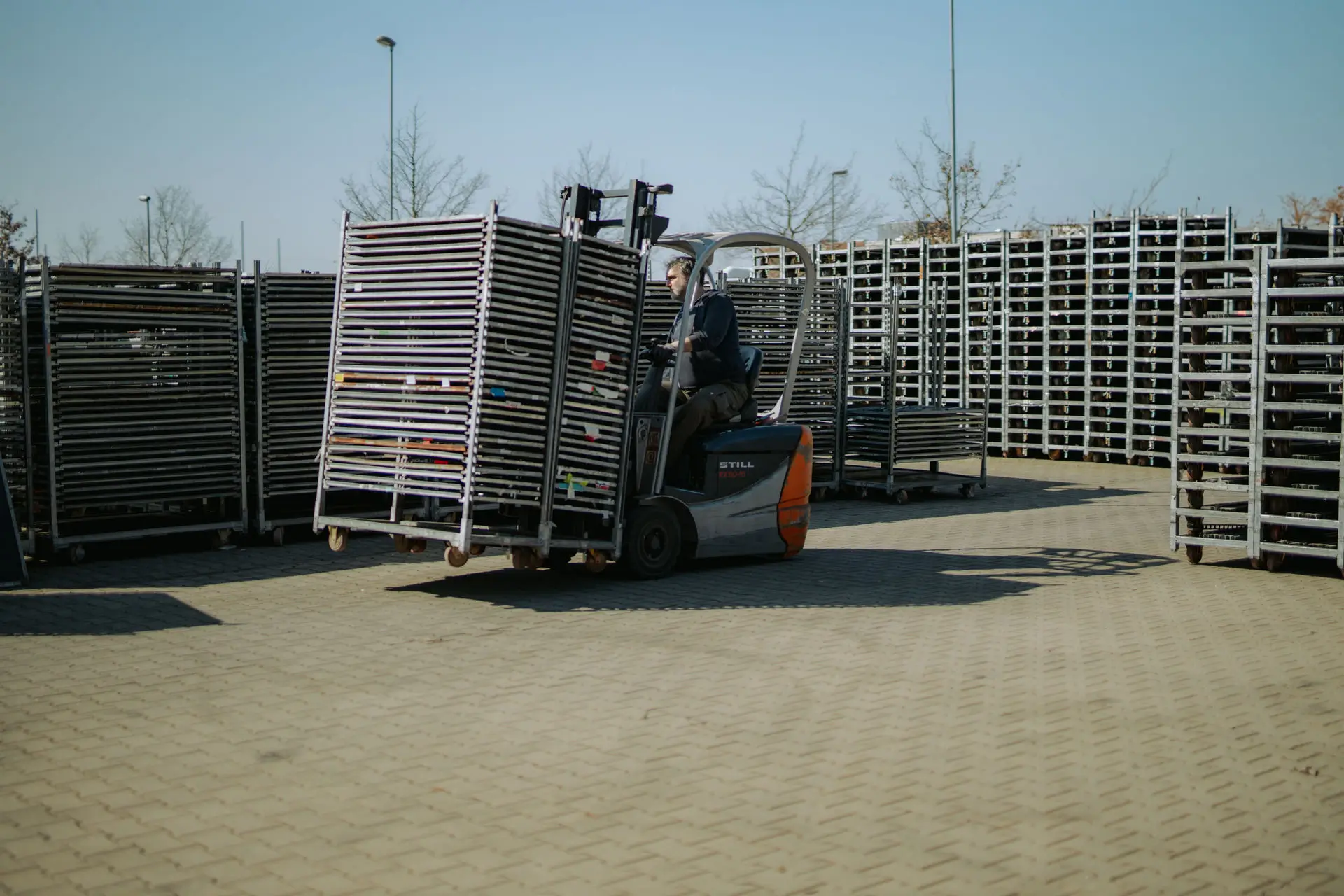 A person is driving a Still forklift truck on a paved outdoor area, transporting stacked metal mesh frames, surrounded by further rows of stored mesh frames.