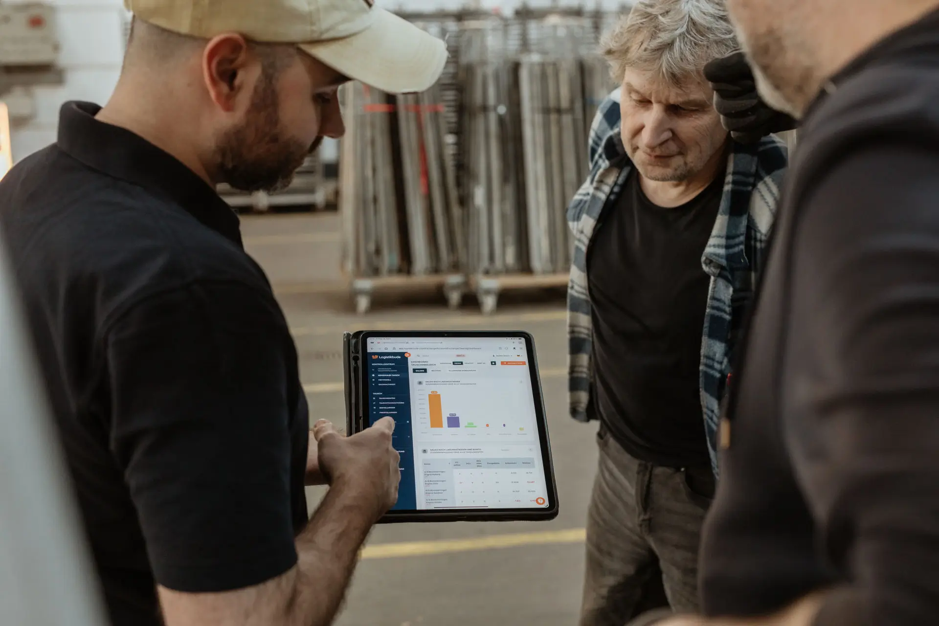 Three people in a warehouse are looking together at a tablet displaying logistics software with charts and overviews.