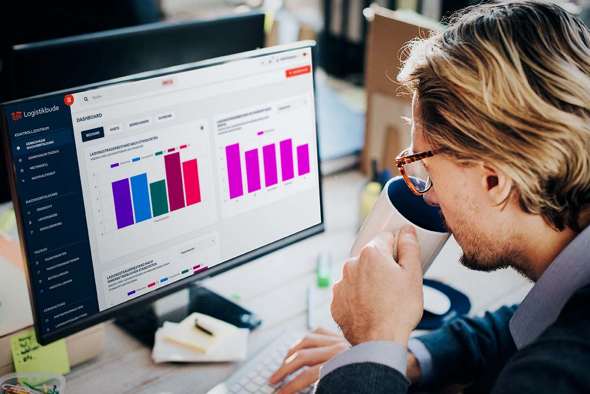 A man wearing glasses drinks coffee while looking at a screen showing the Logistikbude dashboard with colorful bar charts of load carrier inventory.
