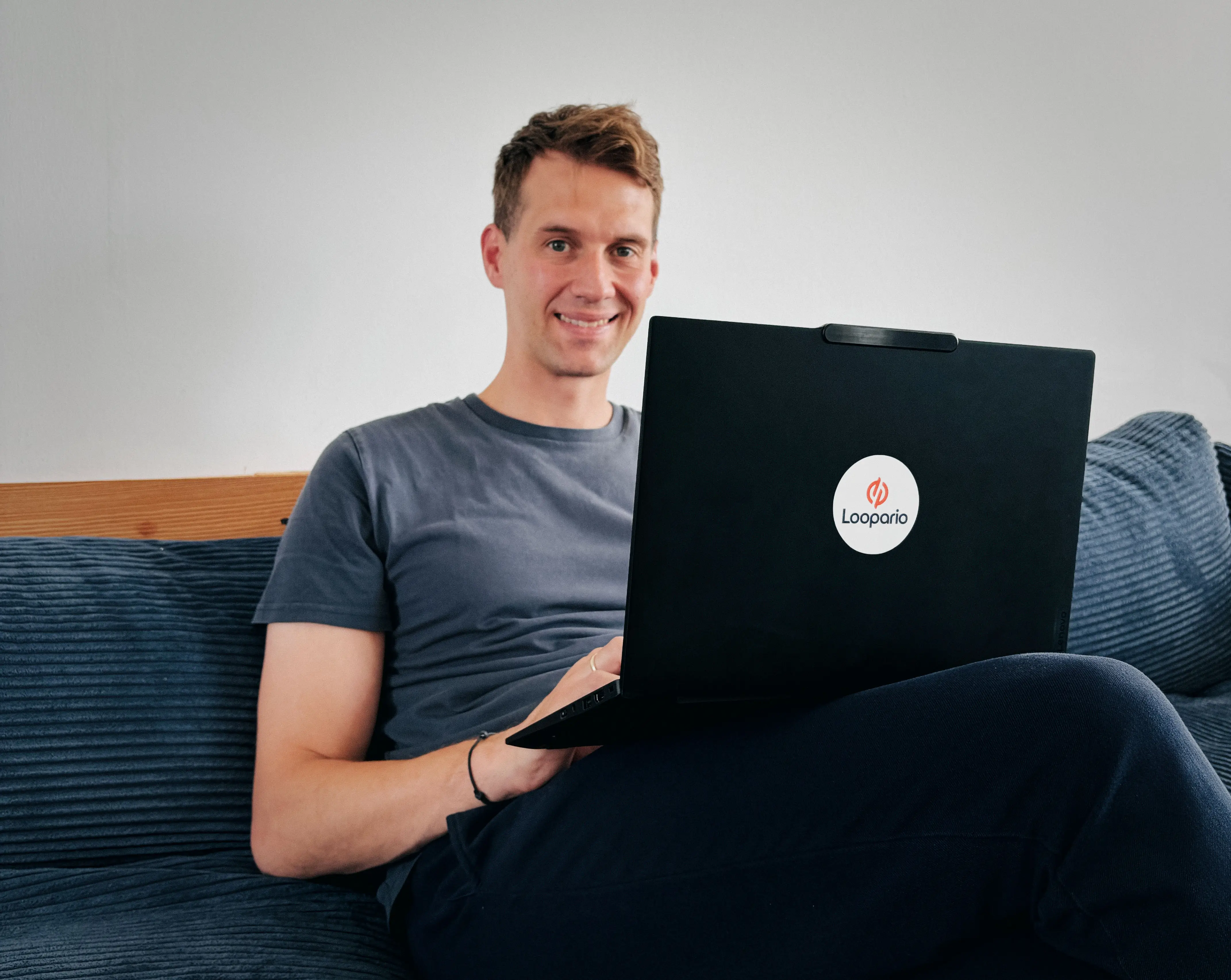 Young man sitting on a blue sofa, presenting a black laptop with the white Loopario logo. He is wearing a dark gray t-shirt and smiling at the camera. The background shows a neutral gray wall and a wooden chair.