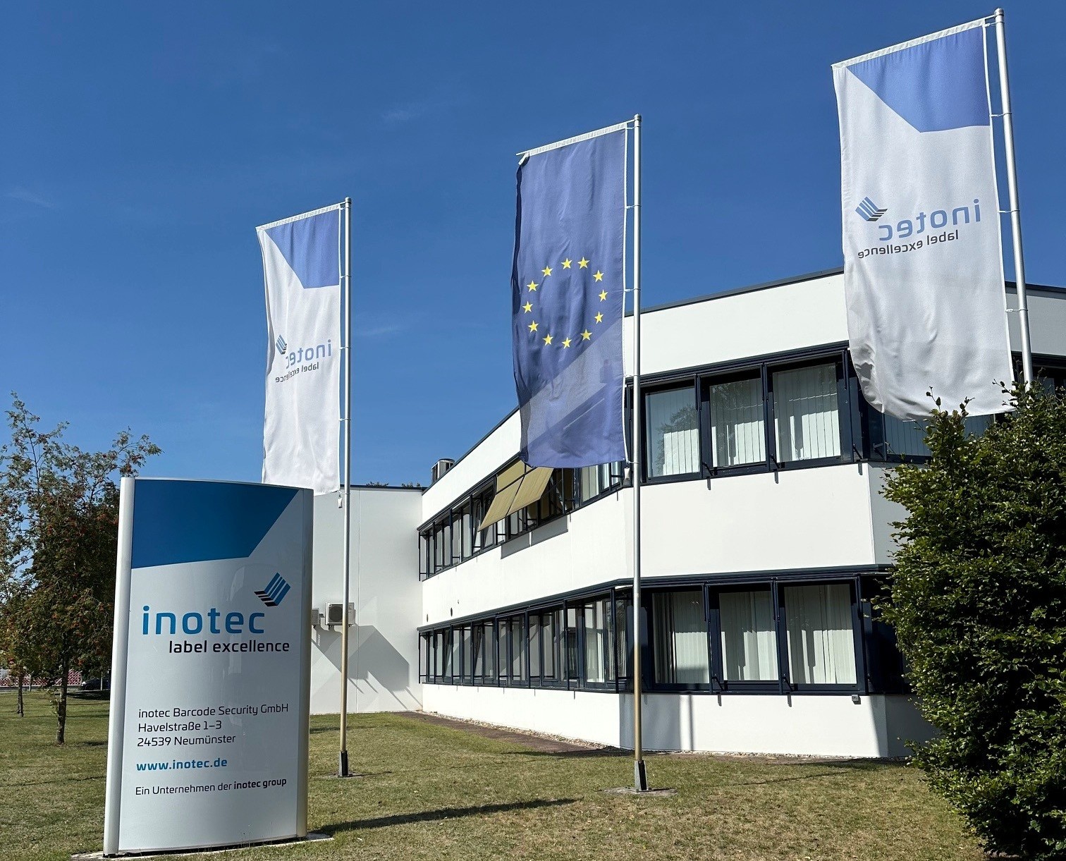 Weißes Bürogebäude der inotec Barcode Security GmbH in Neumünster mit Firmenstele und drei Fahnenmasten (inotec-Fahnen und Europaflagge) vor blauem Himmel.