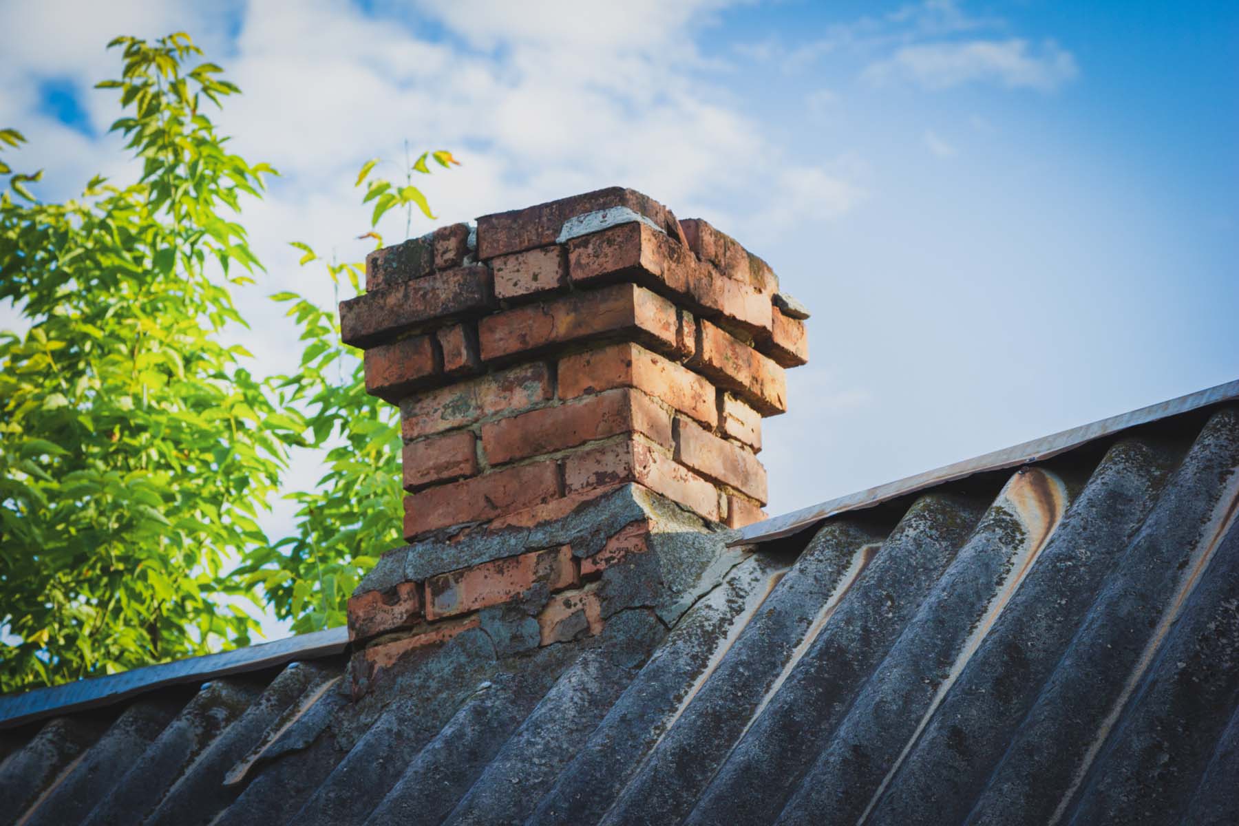 Gammel mursteinpipe på et bølget tak med grønne trær og blå himmel i bakgrunnen.