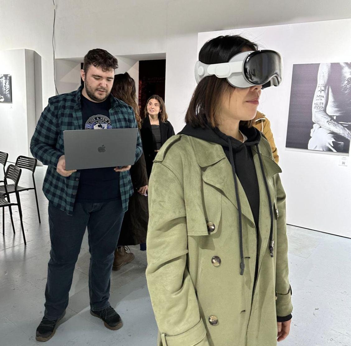 A visitor in a white-walled gallery wearing Vision Pro while a colleague holds a laptop.