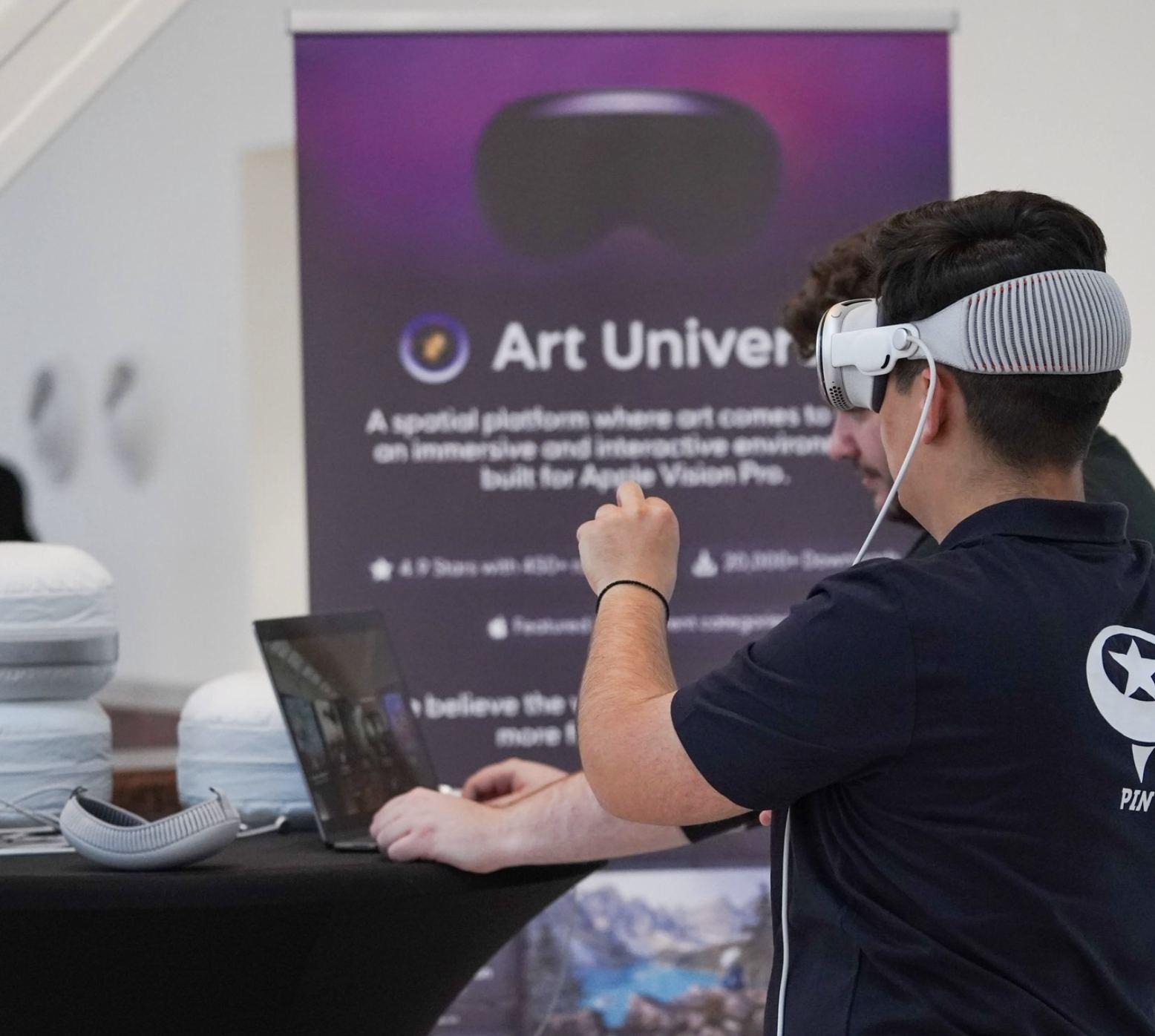 An engineer wearing Vision Pro at a demo table with a laptop, beside the Art Universe banner.