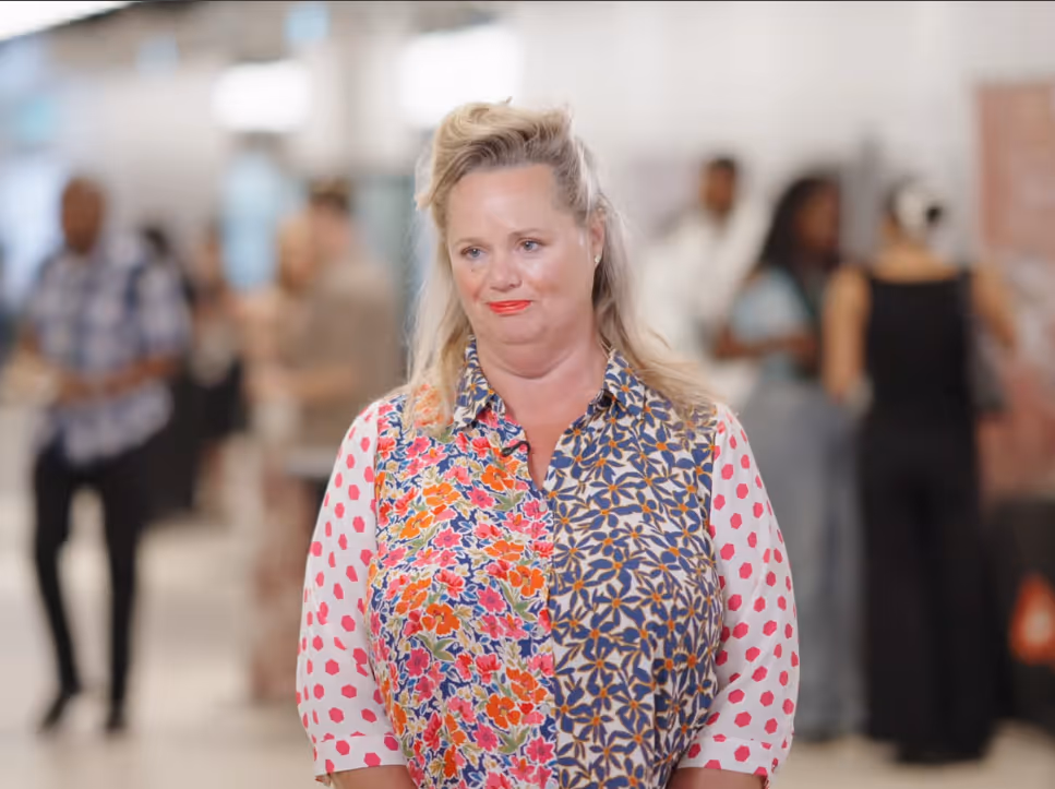 Woman with blonde hair wearing a floral and polka dot patterned blouse standing in a busy indoor environment.