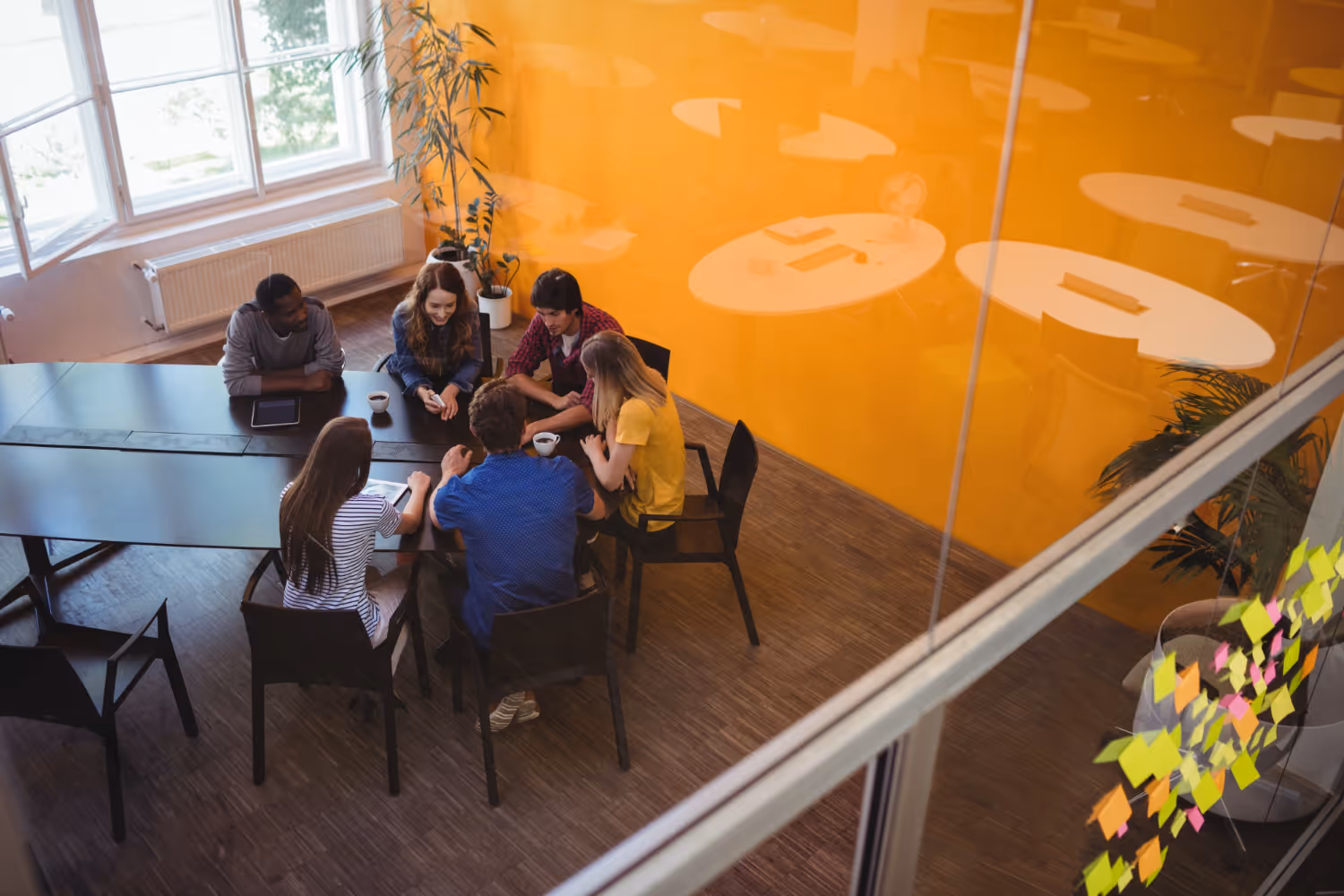 Group of six people sitting around a table in a modern office with a bright orange wall, collaborating and discussing ideas.