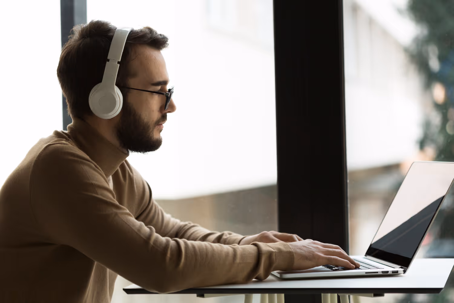 Man wearing white headphones and glasses typing on a laptop by a window.