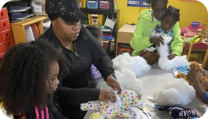 A group of children and adults sit around a table stuffing toy animals with synthetic stuffing in a colorful room.