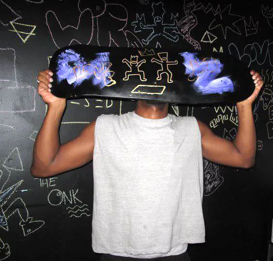 Person holding a skateboard with cartoonish yellow drawings in front of their face against a black wall covered in various chalk drawings.