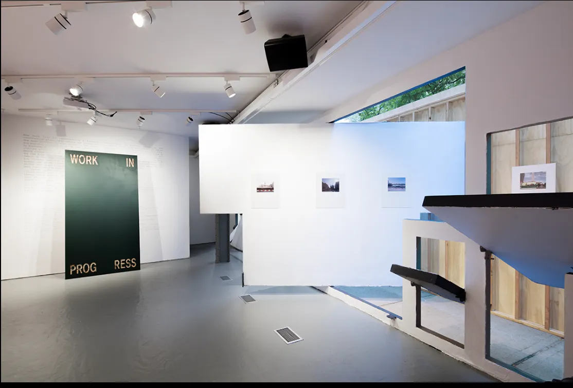 Modern gallery space with white walls displaying small framed photographs and a large piece with the words 'WORK IN PROGRESS'.