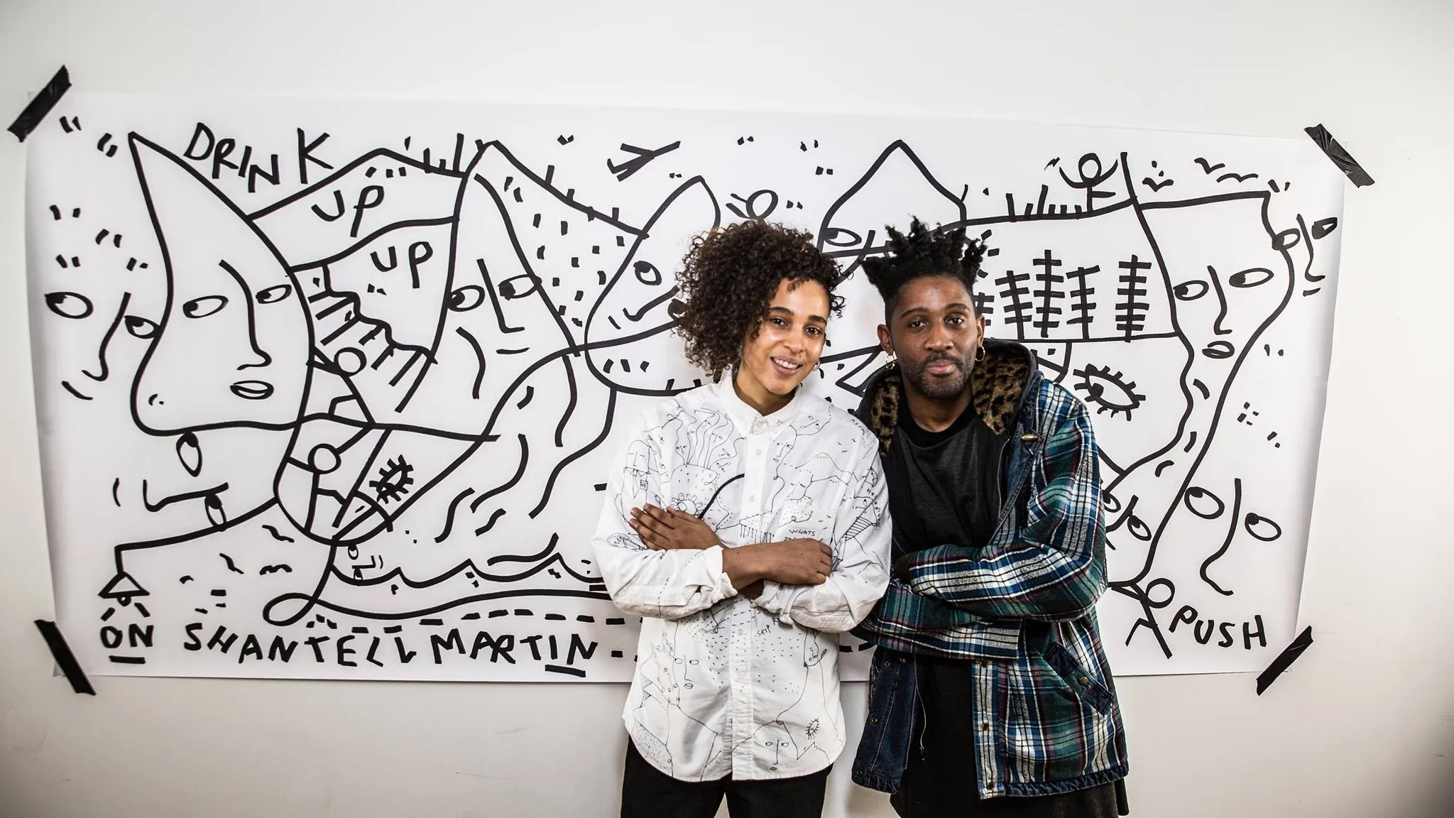 Two people standing with arms crossed in front of a large white paper featuring black line drawings of abstract faces and words like 'DRINK UP' and 'PUSH'.