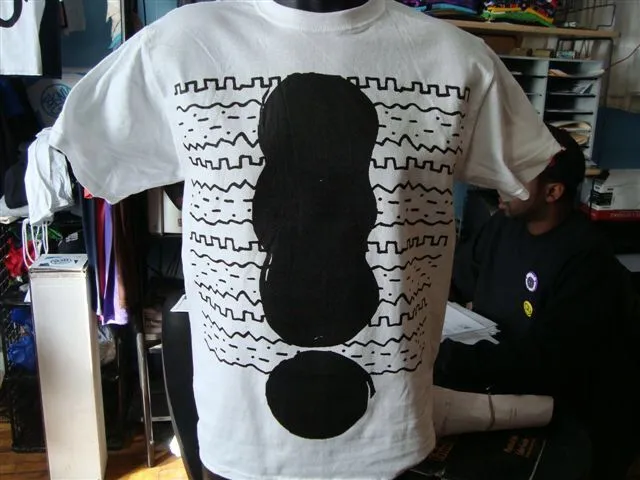 White T-shirt with a large black abstract shape and zigzag lines printed on the front, displayed indoors.
