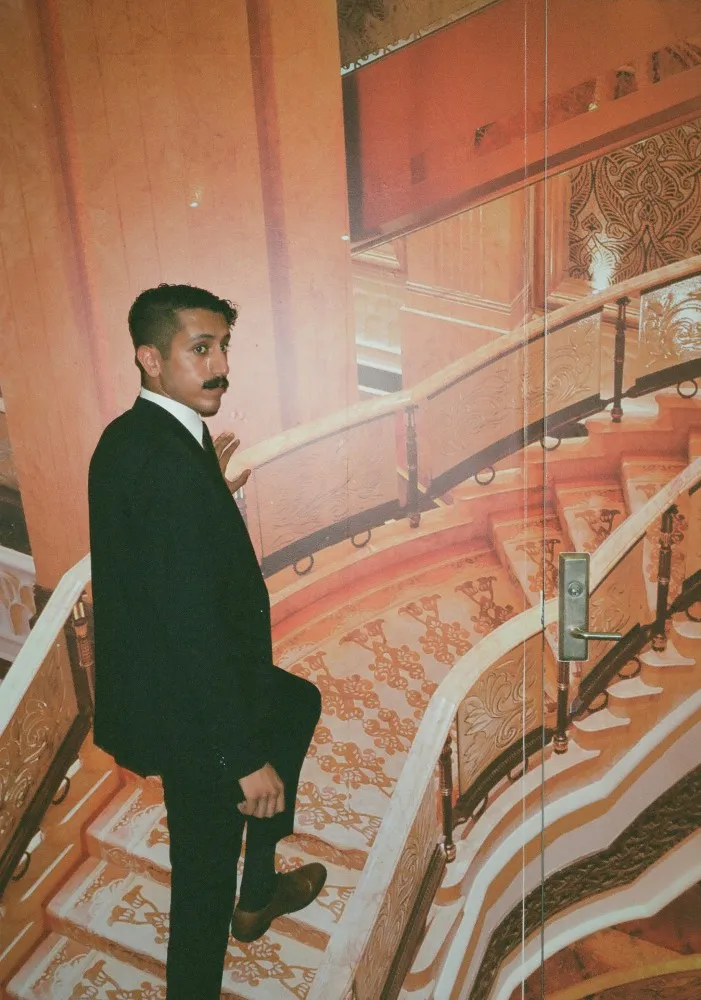 Man with mustache in dark suit ascending ornately patterned staircase with intricate railing.