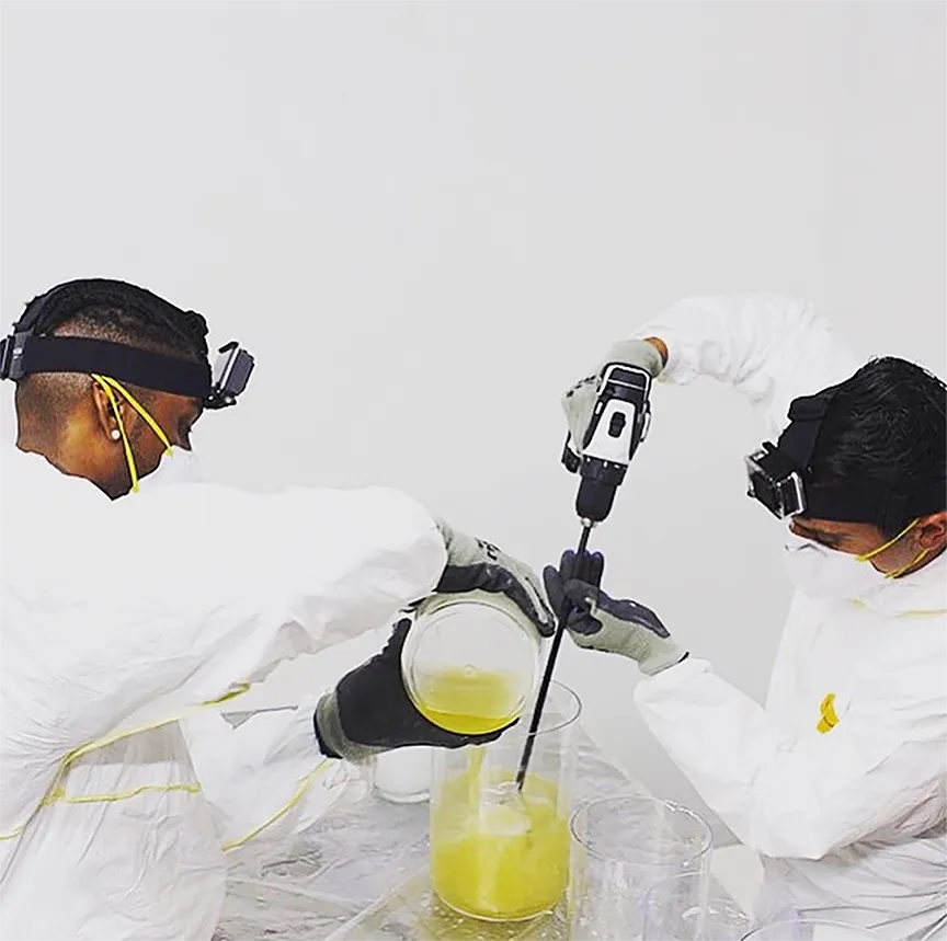 Two people in white protective suits and gloves mixing a yellow liquid with a handheld electric mixer in a clear container.