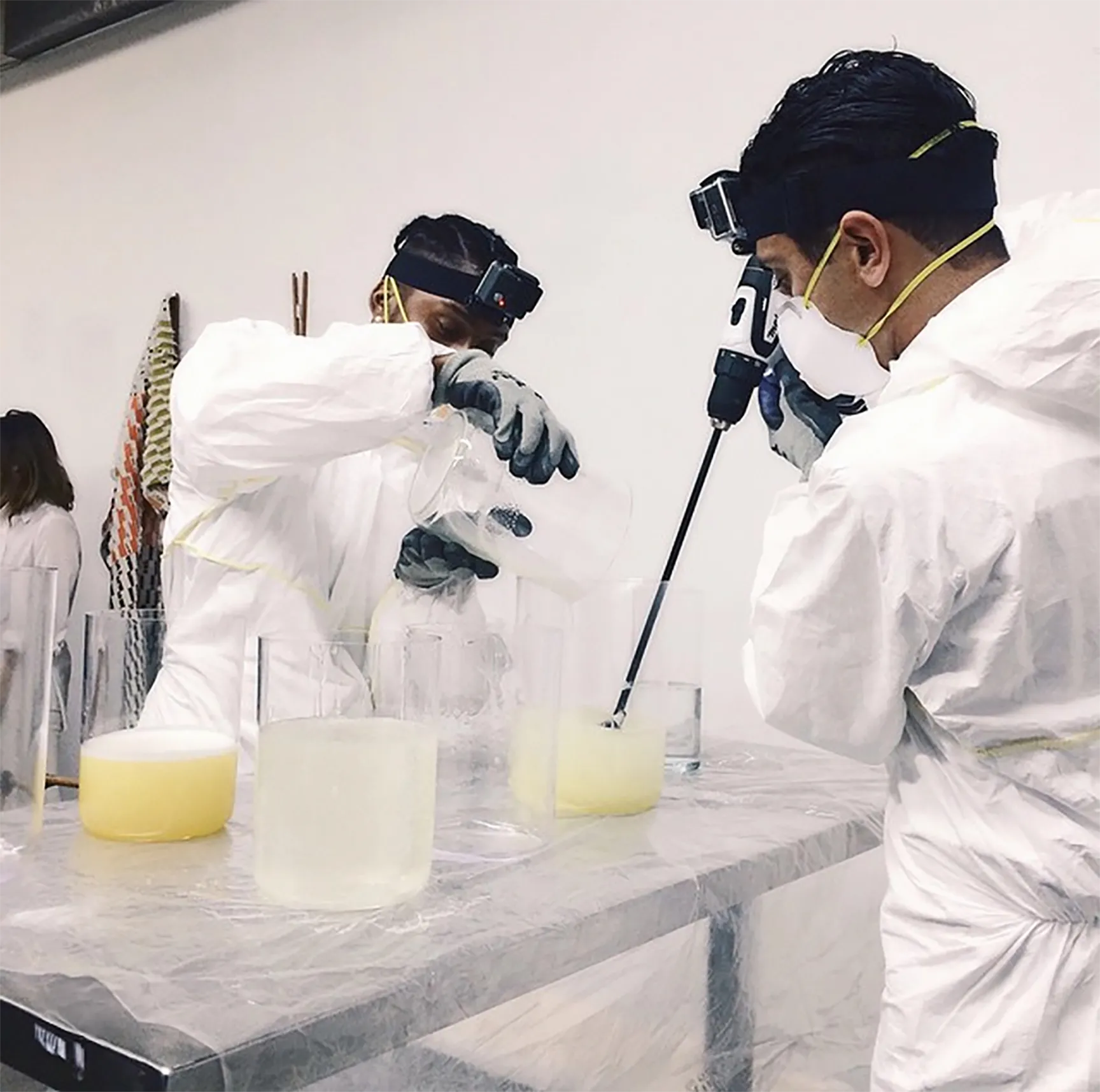 Two people in white protective suits and gloves working with liquid in transparent containers on a table, one pouring liquid and the other using a handheld mixer.