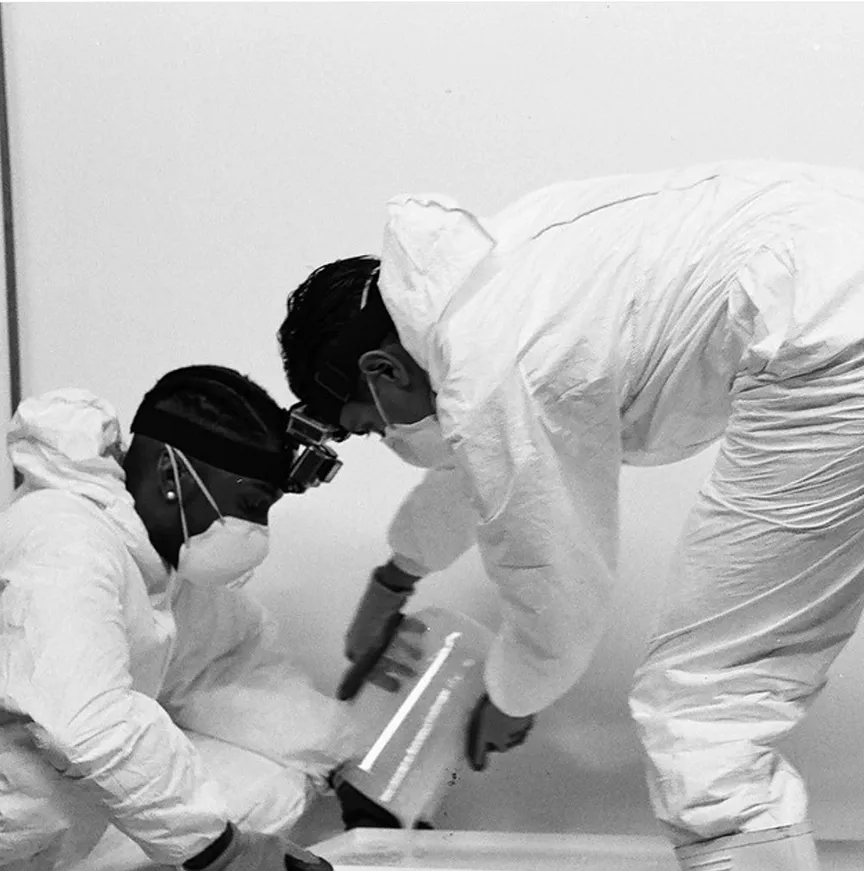 Two individuals in white protective suits and masks working closely, one holding a clear container with white contents.