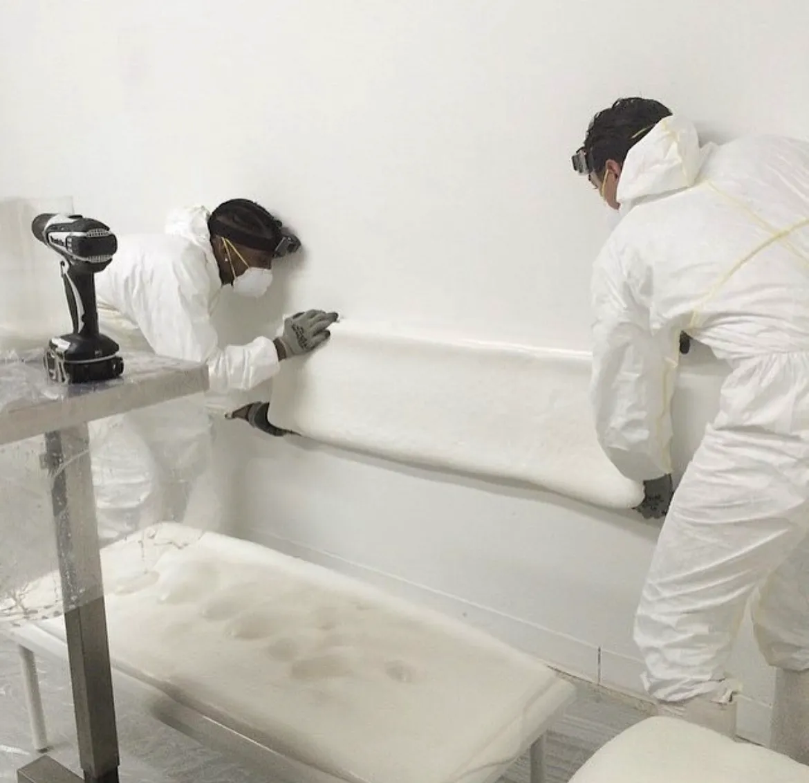 Two workers in white protective suits and masks installing or adjusting a white foam insulation panel on a wall.