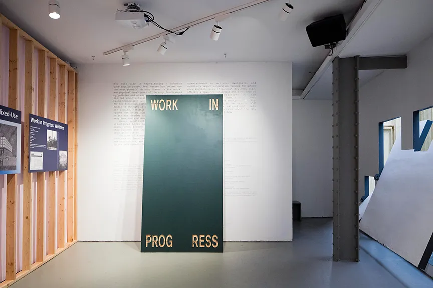 Interior of a modern exhibition space with a large dark green panel displaying the phrase 'WORK IN PROGRESS' in separated letters, mounted against a white wall with text, alongside wooden slats holding information boards.