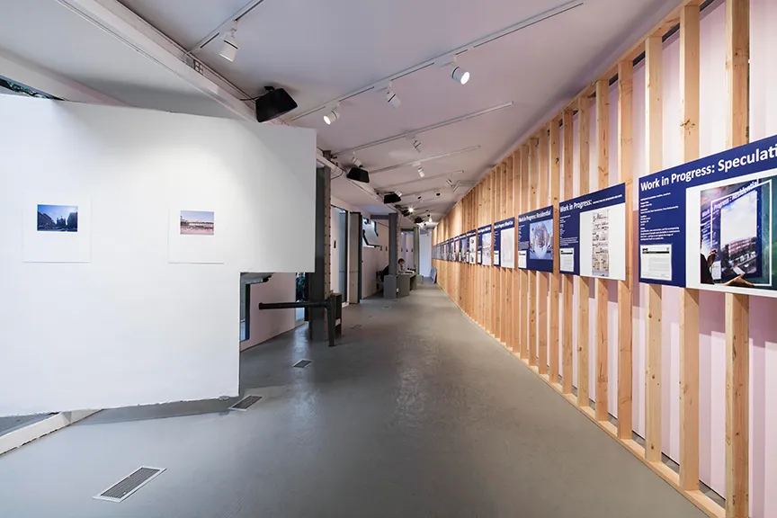 Interior view of a modern exhibition space with wooden slat walls displaying informational panels and two small photographs mounted on a white wall.