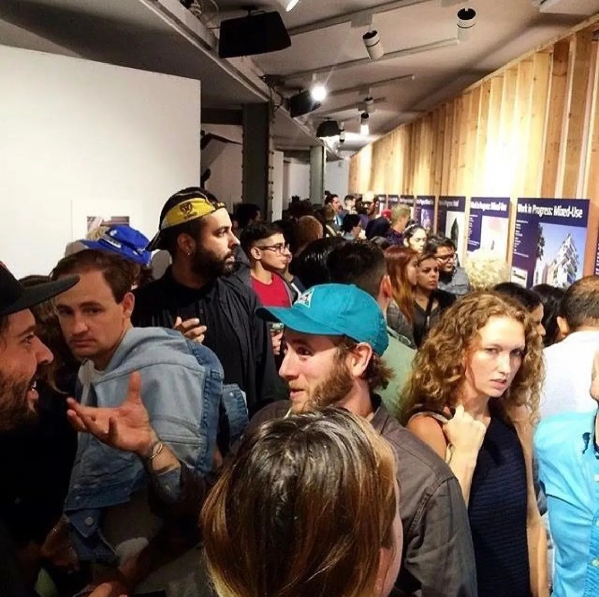 A crowded indoor event with people closely gathered, some engaging in conversation near a wood-paneled wall with informational posters.