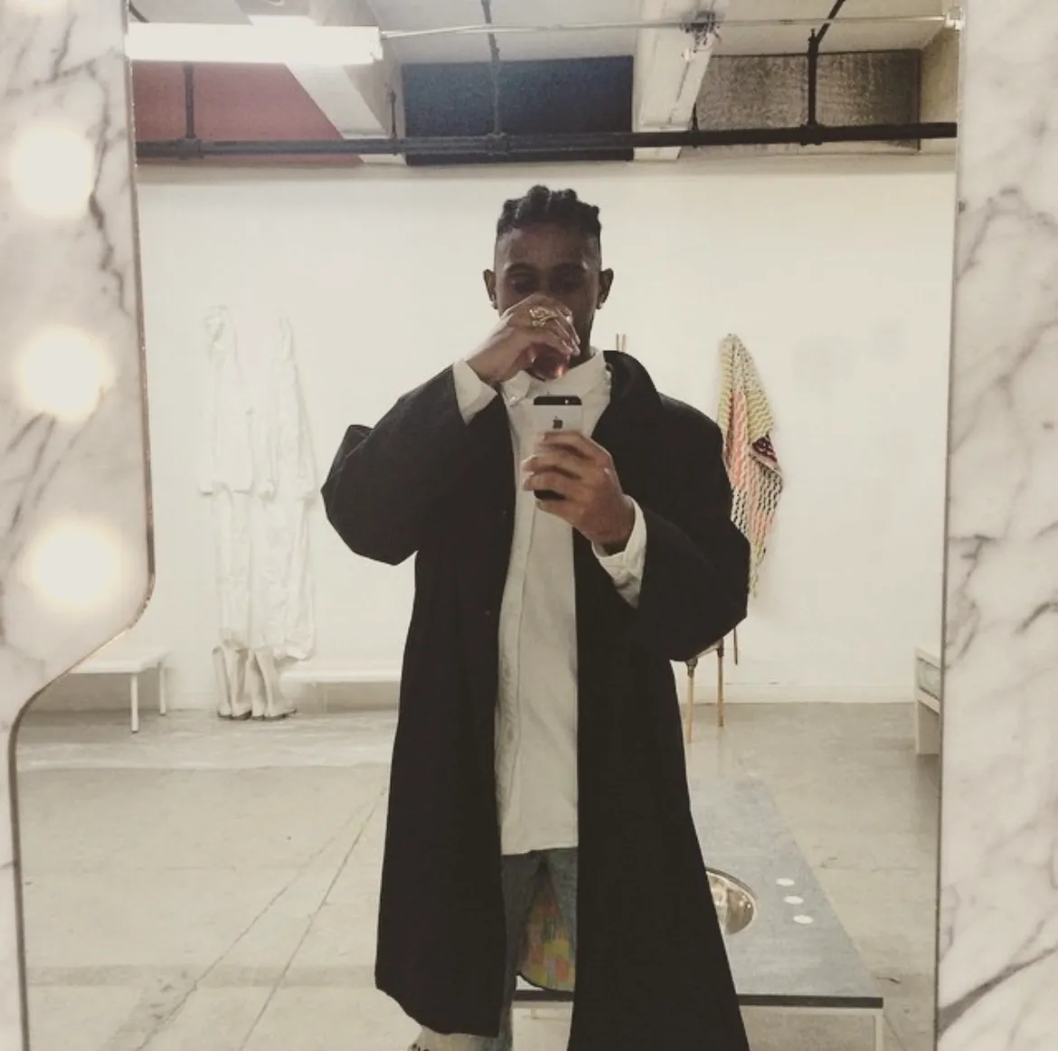Person wearing a black coat and white shirt taking a mirror selfie while drinking from a glass in a minimalist room.