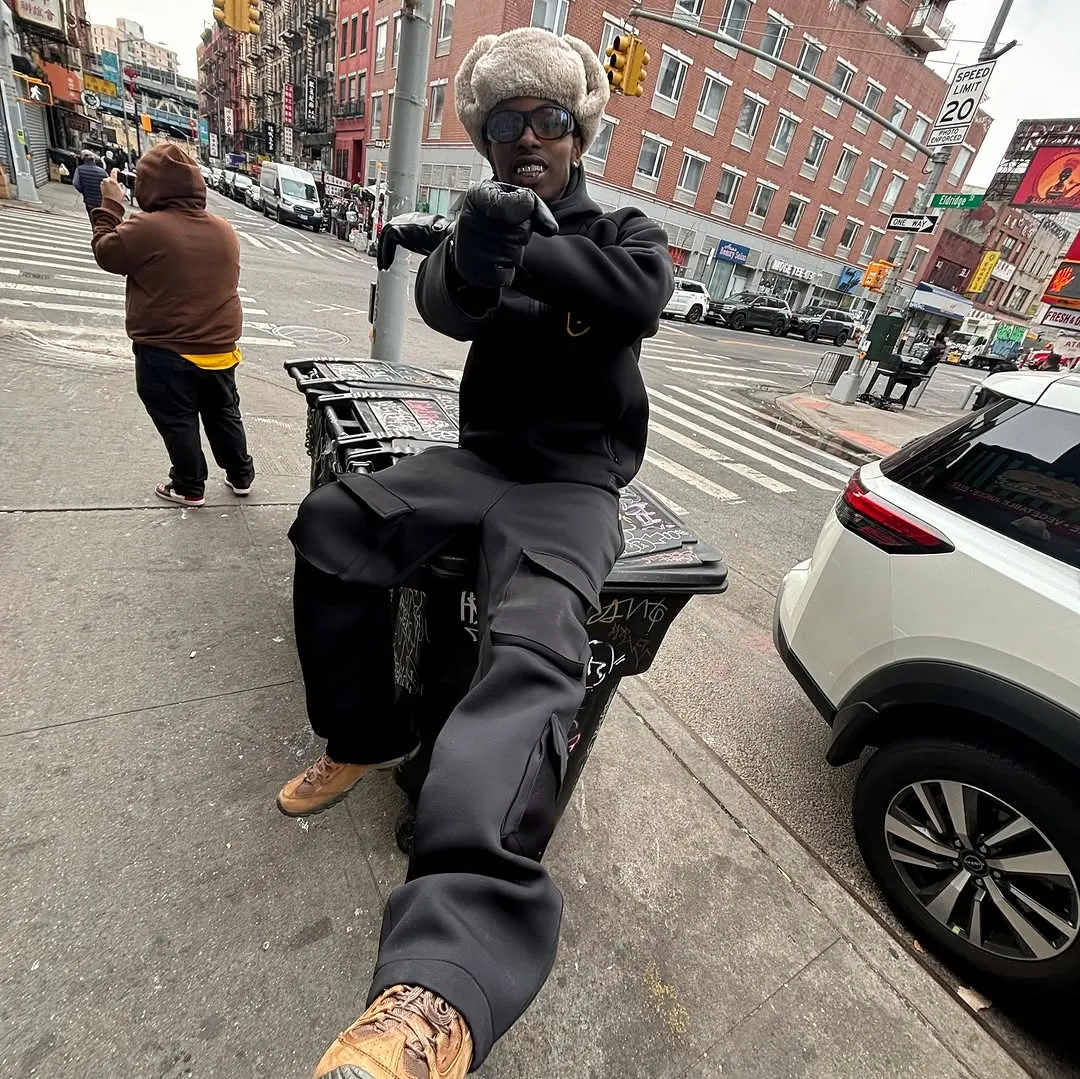 Person wearing a furry hat, sunglasses, and black outfit sitting on a graffiti-covered trash bin, pointing at the camera on a city sidewalk.