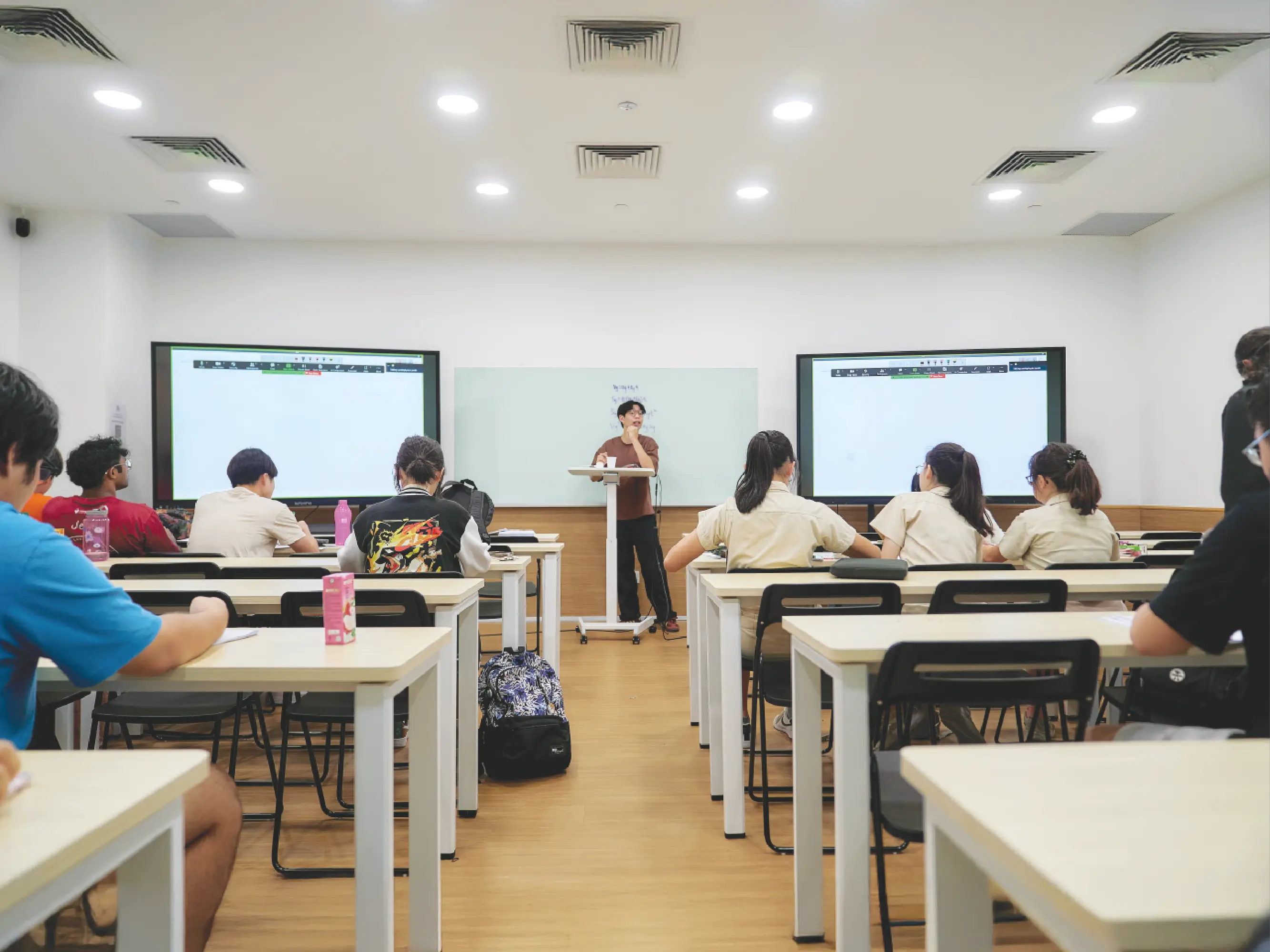 A classroom scene with students seated at desks facing the front, where a person stands at a lectern speaking. Two large screens flank a whiteboard behind the speaker, and the room is brightly lit with ceiling lights. Students are listening, some with notebooks and bags on the floor beside their desks.