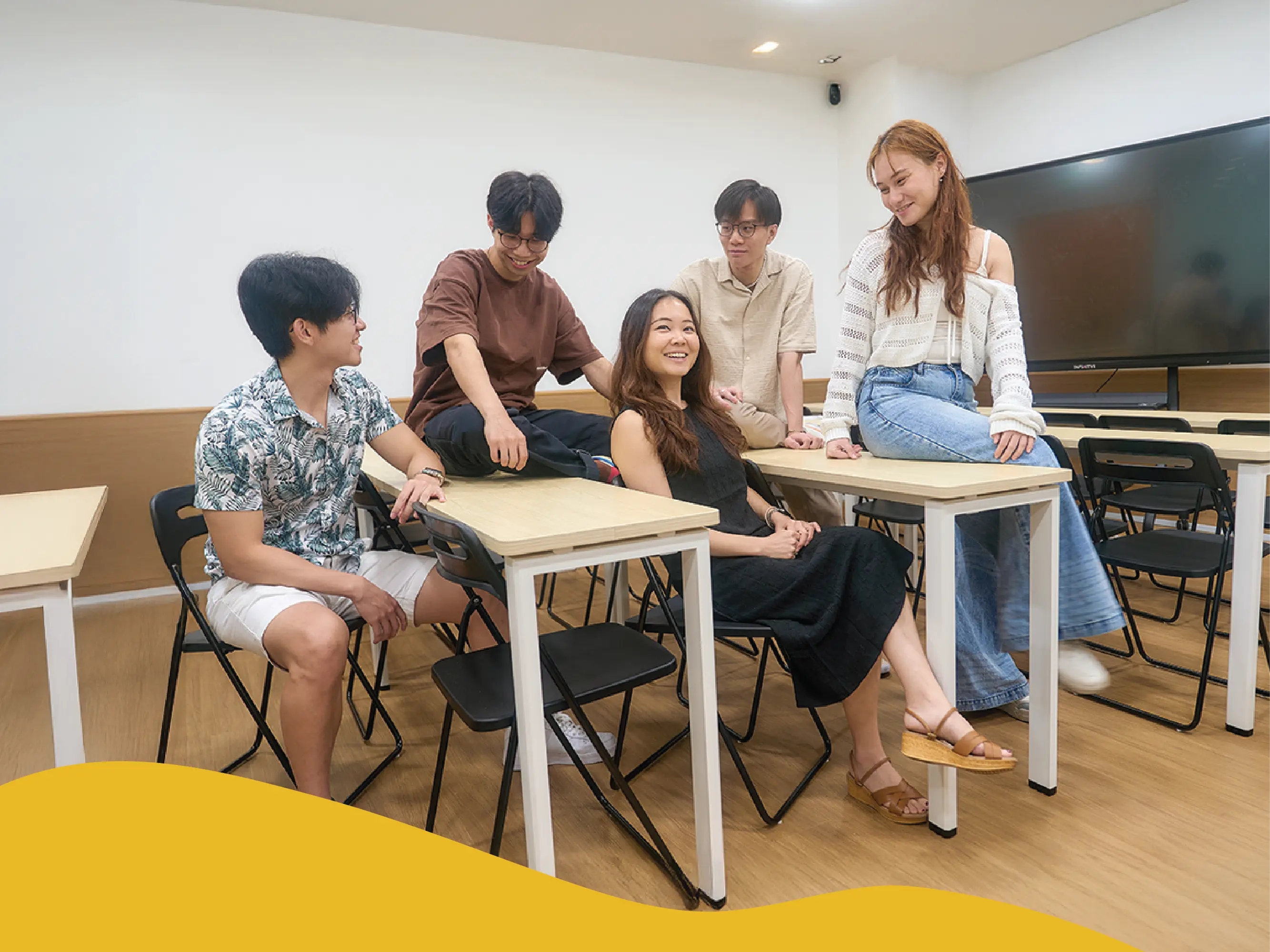 Five students in a modern classroom, seated and leaning on desks in a relaxed group discussion.
