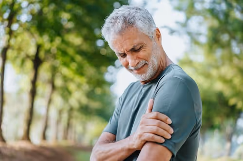 Bildet viser en eldre mann som står i en skog og holder seg til ve overarm i tilsynelatende smerte. Bildet tyder på at mannen har smerter i skulderen, kanskje relatert til rotator cuff tendinopati eller slimposebetennelse eller bursitt i skulderen. 