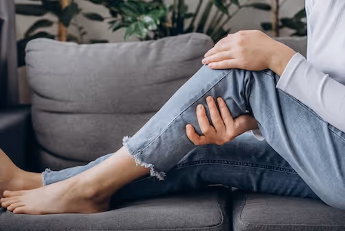 en kvinne sitter på en sofa med bøyd kne og holder seg til oversiden av kneet, knehasen og øverst på bakside av legg, og hinter om at hun har smerter i både foran på kneet, i knehasen eller i leggen. Hun har kanskje betennelse i kneet, eller bakers cyste. 