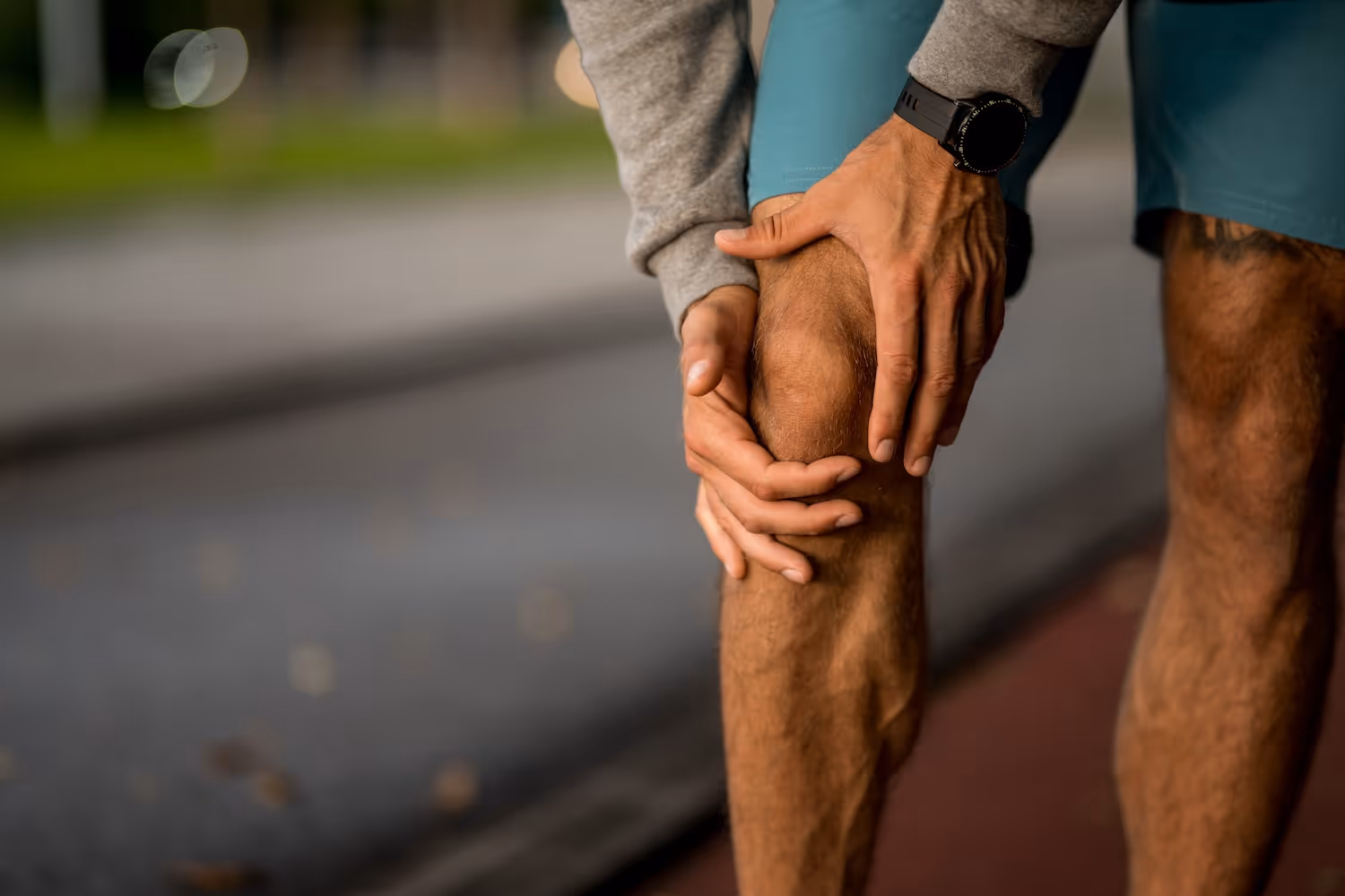 En mannlig løper holder rundt framsiden av kneet på grunn av smerte etter trening eller løping utendørs. Bildet illustrerer knesmerter, mulig patellofemoralt smertesyndrom (løperkne) eller annen belastningsskade. Knesmerter årsaker, behandling og forebygging.