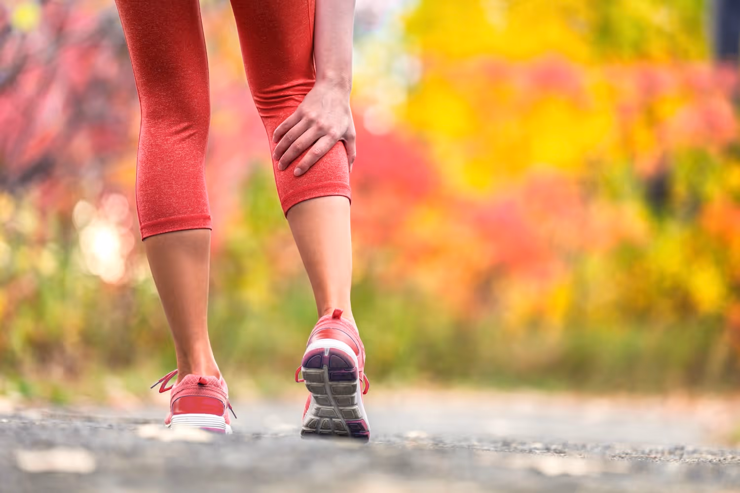 bildet viser en kvinne som løper på en vei med fargerike høstblader på trærne som tar seg bak på leggen på grunn av smerter. 