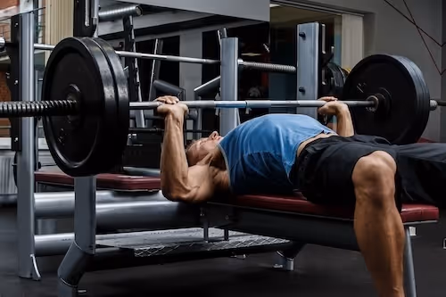 bildet viser en ung muskuløs mann som ligger på et benkpressapparat og løfter benkpress. Bildet viser en typisk aktivitet som belaster acromioklavikular leddet mye, og en typisk årsak til AC-ledds smerter relatert til forstuing av AC-leddet.