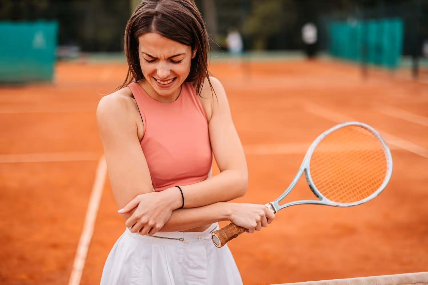 Bildet viser en kvinne på en grusbane som holder seg til utsiden av albuen med smerte etter å ha spilt tennis. Hun viser tegn til tennisalbue (lateral epikondylitt), en vanlig belastningsskade i albuen blant tennisspillere.