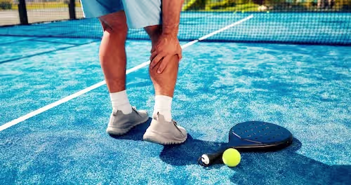 En padelspiller står på banen og holder seg fast i baksiden av leggen med et smertefullt uttrykk, som illustrasjon på en akutt strekk i leggmuskulaturen (tennislegg). En padelracket og ball ligger på det blå dekket i forgrunnen.