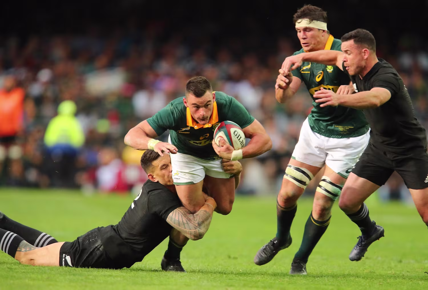 Rugbyspiller i grønn drakt løper med ball og blir taklet lavt av motstander i svart drakt — illustrasjonsbilde av kontaktidrett og typisk skademekanisme for kneskader som MCL-skade.