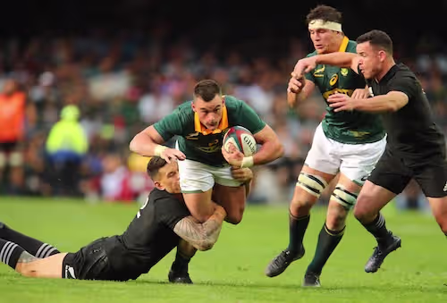 Rugbyspiller i grønn drakt løper med ball og blir taklet lavt av motstander i svart drakt — illustrasjonsbilde av kontaktidrett og typisk skademekanisme for kneskader som MCL-skade.