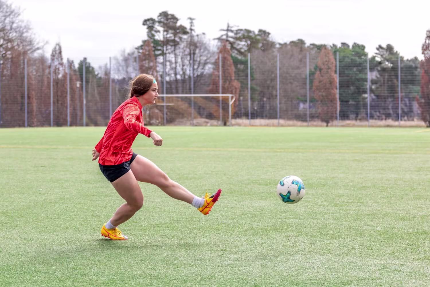 Kvinnelig fotballspiller som sparker ballen på kunstgress – illustrasjon av spark, en vanlig årsak til lyskestrekk (strekk i adduktorene på innsiden av låret).