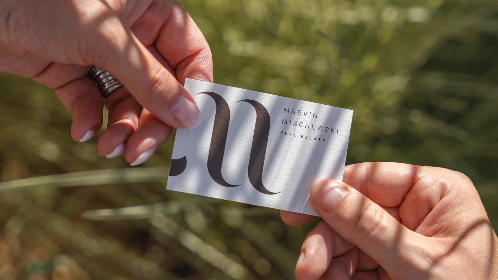 Close-up of two people holding a business card outdoors; the card features a large stylized “M” and the text “Marvin Mischewski — Real Estate” against a blurred green background.