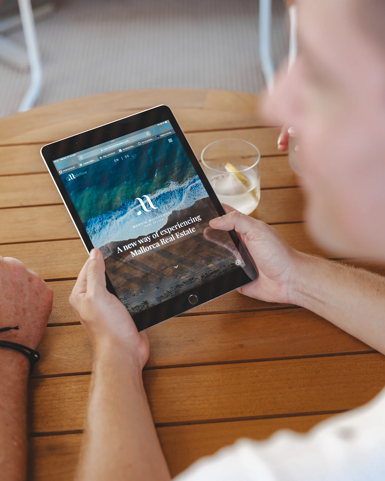 Close-up of a person holding a tablet at a café table, showing Marvin Mischewski real estate website with a coastal image; a lemon water sits beside it.