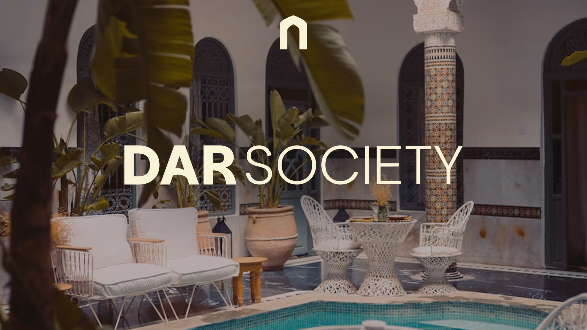 Ornate courtyard with a small turquoise pool, surrounded by white wicker chairs, potted plants, intricate tilework, and Dar society logo