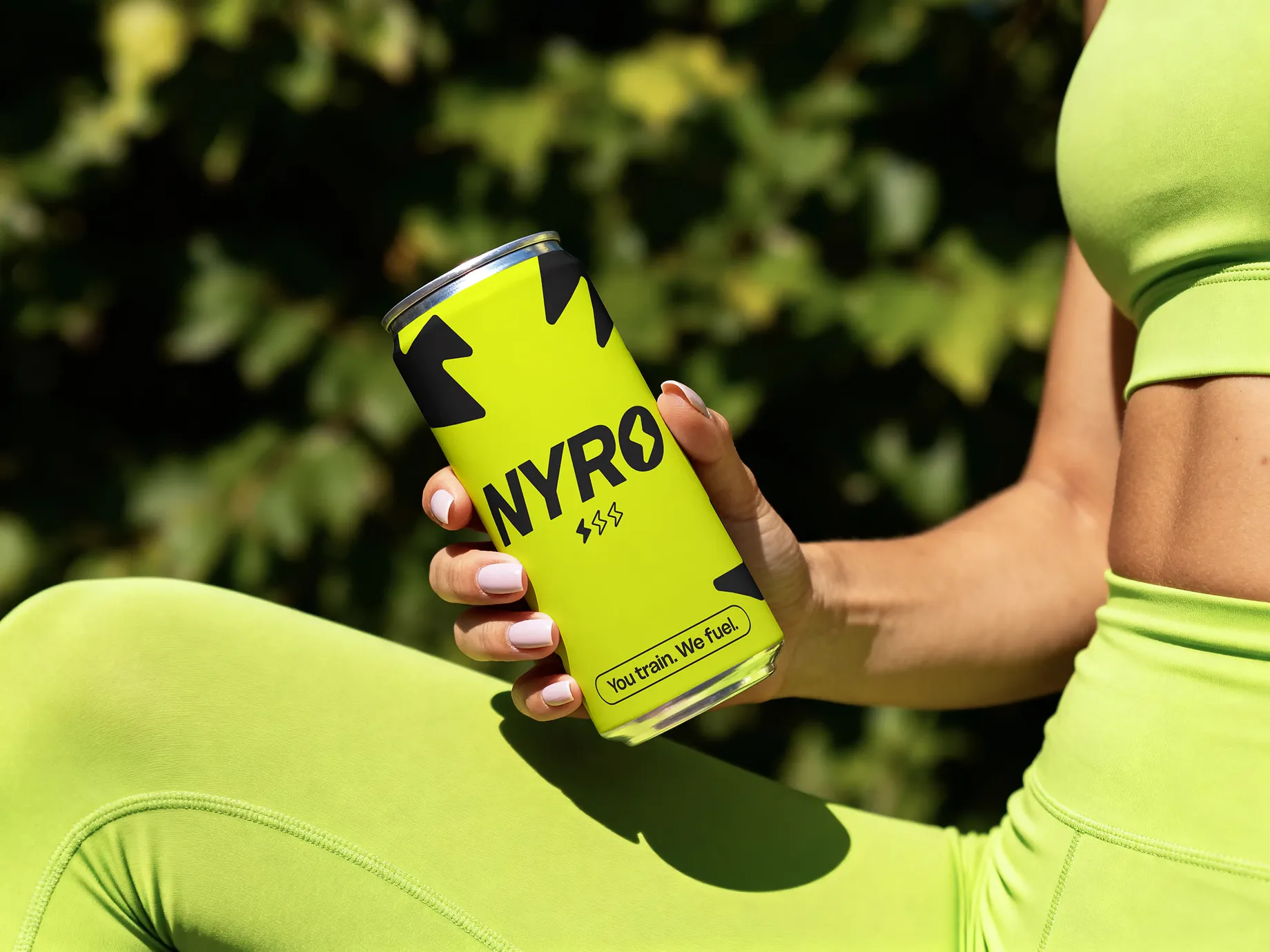 A person wearing lime green athletic wear holds a yellow-green can labeled NYRO with black lightning bolt designs and the tagline 'You train. We fuel.' outdoors with blurred greenery in the background.