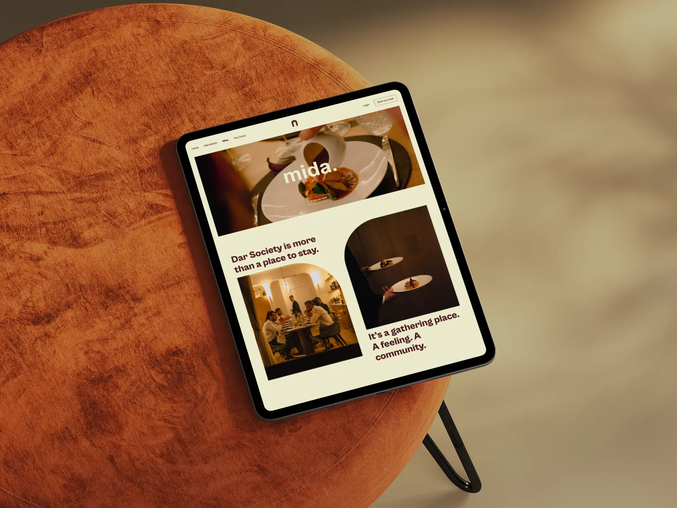 Tablet on a round rust-colored ottoman displaying a website for 'mida,' featuring images of a plated dish being served and people dining together, with text about Dar Society being more than a place to stay and a sense of community.