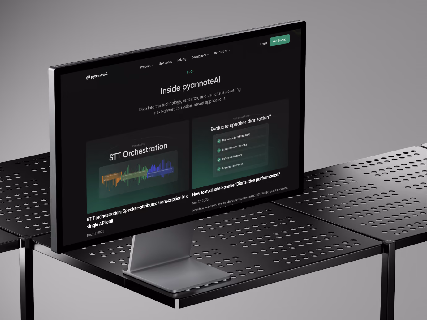 A modern flat-screen monitor on a perforated black desk displaying the pyannoteAI blog homepage with sections on STT Orchestration and evaluating speaker diarization performance.