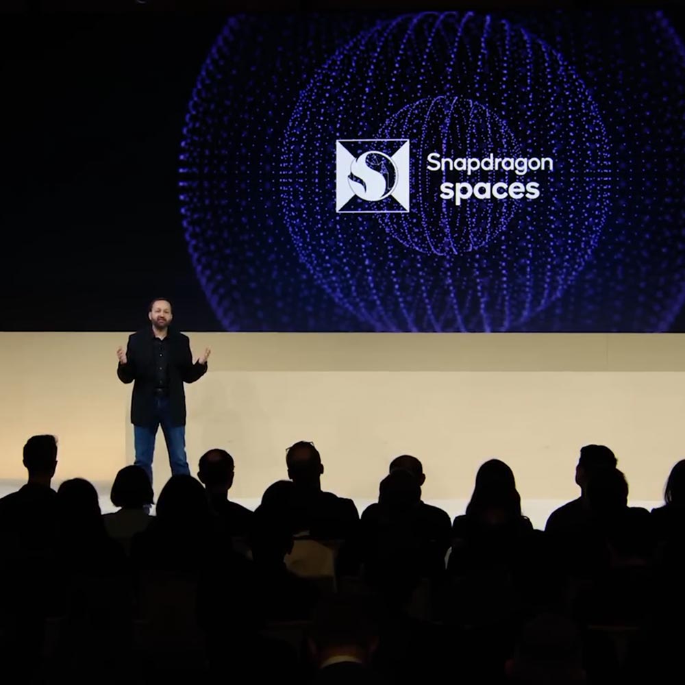 A male speaker stands onstage at the XR Unlocked event in front of a screen that reads "Snapdragon spaces."