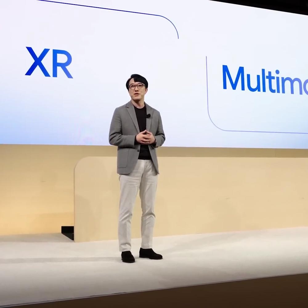 A male speaker stands onstage at the XR Unlocked event in front of a screen that reads "XR" and "Multimodal AI" in blue text.